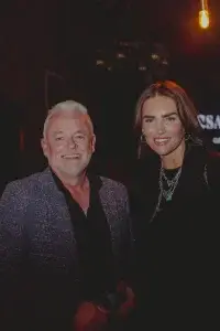 Smiling white-haired man in a grey blazer beside a woman with long brown hair and chain necklaces in a dimly lit event.