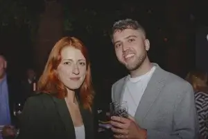 Two smiling people at a night event: red-haired woman in a dark blazer and a bearded man in a grey blazer holding a pint