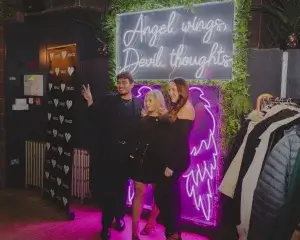 Three people pose in front of a neon sign reading 'Angel wings, Devil thoughts' with glowing purple angel wings and foliage.