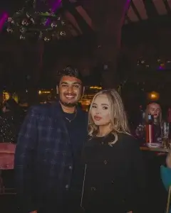 Smiling man in a dark blazer poses with a blonde woman in black at a dimly lit bar with warm lights