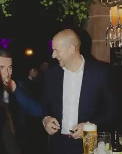 Bald man in a dark suit laughing at a bar, holding small items while a pint of beer and glasses sit in the foreground