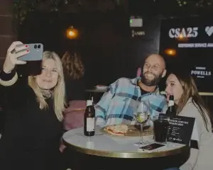 Three people posing for a selfie: woman holding a phone, man in a checked shirt and a brunette, drinks and pizza on table.