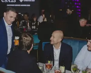 Well-dressed men chatting at a booth in a dimly lit awards-night bar, one holding a pint and another smiling at the table.