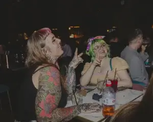 Two women laughing at bar: one with floral sleeve tattoos and pink fringe, the other with green hair, glasses and a bandana.