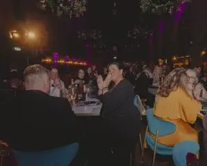 Woman in a black jacket clapping at a crowded, dimly lit restaurant table with guests, wine glasses and purple uplighting.