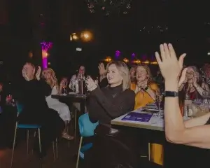 Audience applauding in a dimly lit venue, woman with short blonde hair in dark top smiling and clapping.
