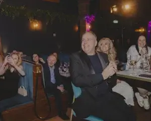 Smiling man in a black blazer clapping at a dimly lit restaurant table, a blonde woman smiling beside him and guests nearby.