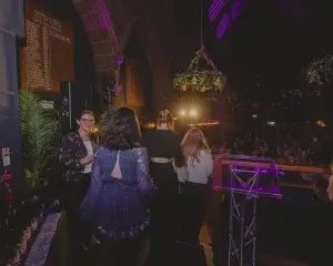 Women on a decorated stage facing an applauding audience in a dim, purple-lit hall with a lectern.