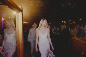 Blonde woman in a white strappy dress with a thigh slit walking through a dim, warmly lit venue as guests watch and clap.