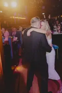 Couple embracing—woman in white dress smiling as man in dark suit holds her, audience clapping in dim venue.