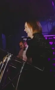 Laughing woman at a podium in profile with short brown hair and zigzag-patterned black dress, purple-lit stage and a blurred man behind