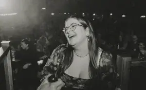 Woman with glasses laughing in a dim, crowded venue, wearing a floral dress and beaded necklace.