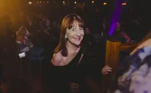 Smiling woman in a black off-shoulder dress with a silver strap, leaning on a wooden railing in a dim crowded venue.