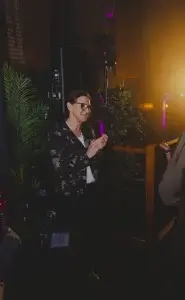 Middle-aged woman in glasses and a dark floral blazer clapping her hands beside stage plants under warm amber lighting.