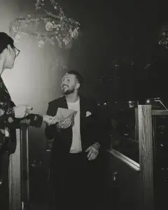 Smiling bearded man in a blazer receiving an envelope from another person in a dim, decorated venue with hanging bulbs