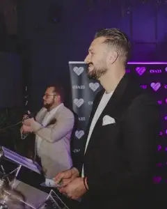 Man in black blazer with white pocket square smiling in profile at a podium, holding notes while a second man claps before a heart-logo event backdrop.
