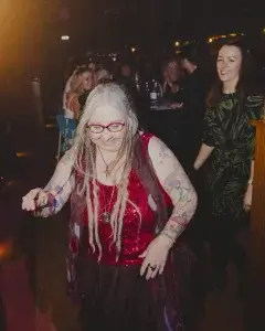 Older woman with long grey dreadlocks, pink glasses and a red sequin top dancing at a dimly lit party with tattooed arms.