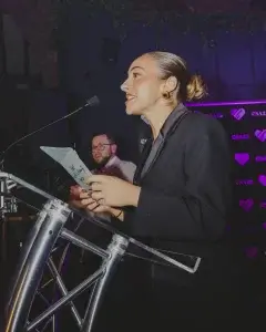 Woman in a black blazer with hair in a bun and gold square earrings speaking at a podium, holding notes with a purple heart logo backdrop.