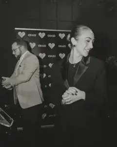 Woman in dark blazer, hair in a bun, smiling with clasped hands before a CSA25 heart-logo backdrop.