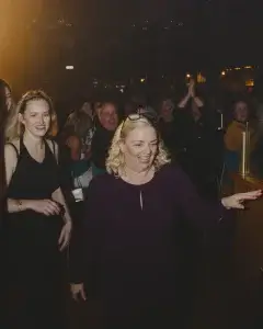 Blonde woman in a dark purple dress with glasses on her head laughing and reaching forward as a crowd applauds in a dimly lit party