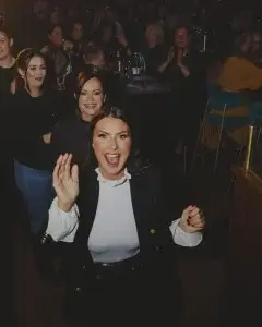 Smiling woman in a white ruffled blouse and black blazer waving excitedly towards the camera in a dim, crowded venue.
