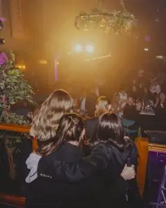 Three women seen from behind with arms around each other, watching a brightly lit stage and applauding audience in a club.