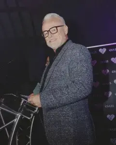 Older man with white hair, beard and glasses in a grey patterned blazer smiling at a podium while holding notes.