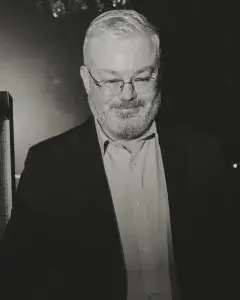 Older man with short white hair and beard, wearing glasses and a dark jacket, smiling and looking down.