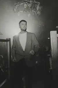 Young man in blazer and white t-shirt walking through a dim, smoky club under hanging bulbs and foliage, black-and-white.