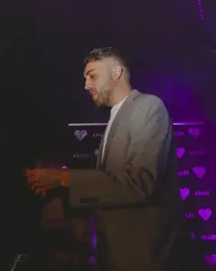 Man in grey blazer with trimmed beard under purple stage lights holding a card before a heart-logo backdrop.