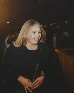 Smiling blonde woman in a black outfit with a gold chain purse strap, seated in a dimly lit venue with hands clasped.