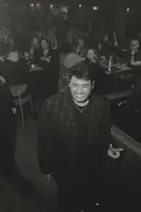 Man in a dark plaid jacket with eyes closed and a pained grin, gesturing in a crowded dim club.