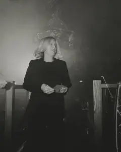 Woman in a black suit with blonde hair smiling and holding a card on a dim, smoky stage under a chandelier.