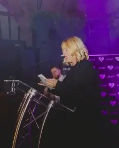 Blonde woman in black reading from notes at a lectern, smiling against a purple-lit backdrop with heart logos.