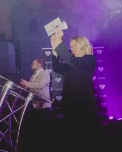 Woman on stage smiling and holding up an envelope toward the audience under purple lighting as a man claps behind her.