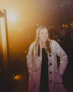Smiling young woman in a white patterned coat standing at a dimly lit evening event with warm backlight behind her.