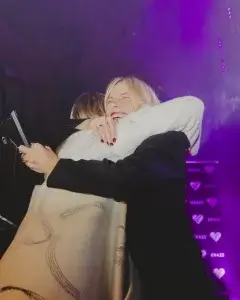 Warm embrace on a purple-lit stage: a smiling blonde with red lipstick hugs someone in a cream patterned shawl with a plaque.