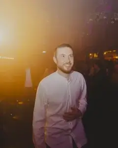 Smiling bearded man in a white button-up shirt bathed in warm orange club lighting with blurred crowd behind him.