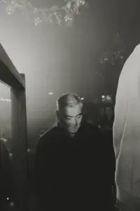Middle-aged man with short grey hair looking down in a dim, black-and-white crowded venue with blurred lights and hanging decorations.