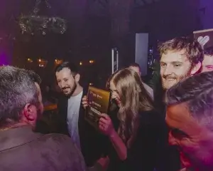 Crowd of smiling adults in a dim venue, a woman laughing and holding a framed award plaque while friends congratulate her.