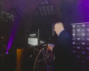 Man in a dark suit speaking at a podium under purple lighting, with an awards slide projected and a CSA25 heart-logo backdrop.