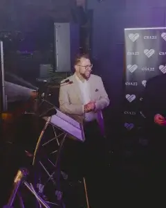 Smiling bearded man in a beige blazer and glasses standing at a lectern with microphone, CSA25 heart-logo backdrop.