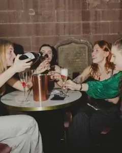 Four women at a round table clinking champagne flutes while one pours from a bottle; raspberries visible in some glasses.