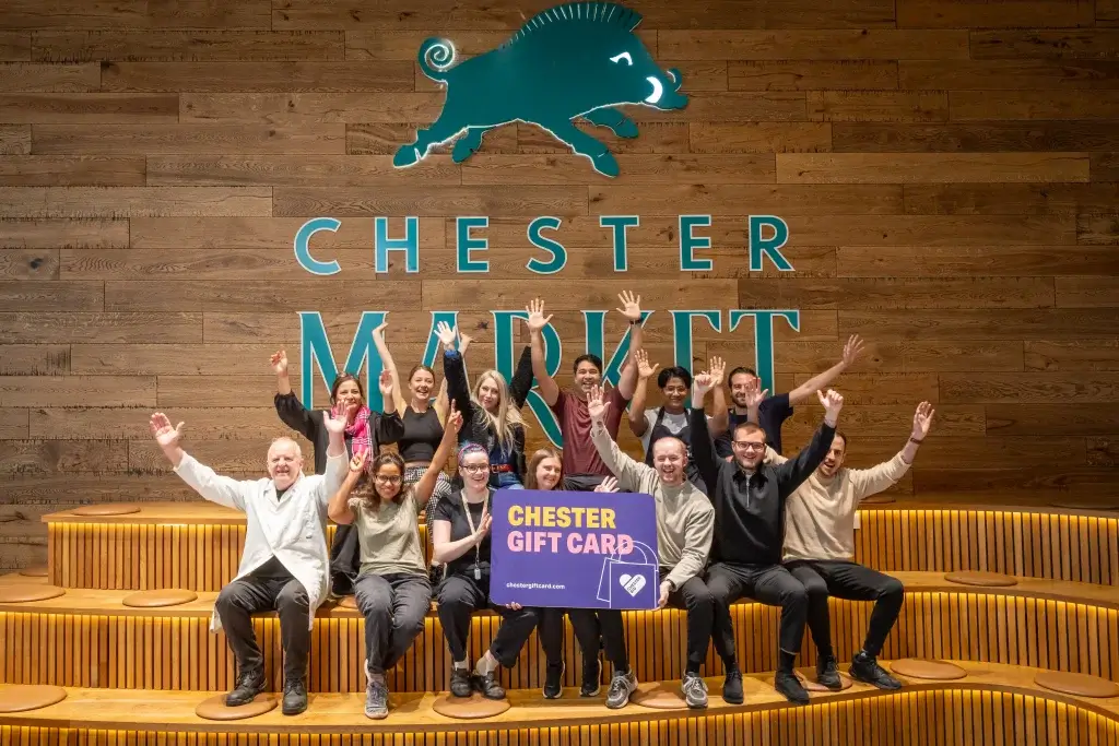 A group of market traders joyfully posing with a Chester Gift Card in front of a "Chester Market" sign on a wooden wall.