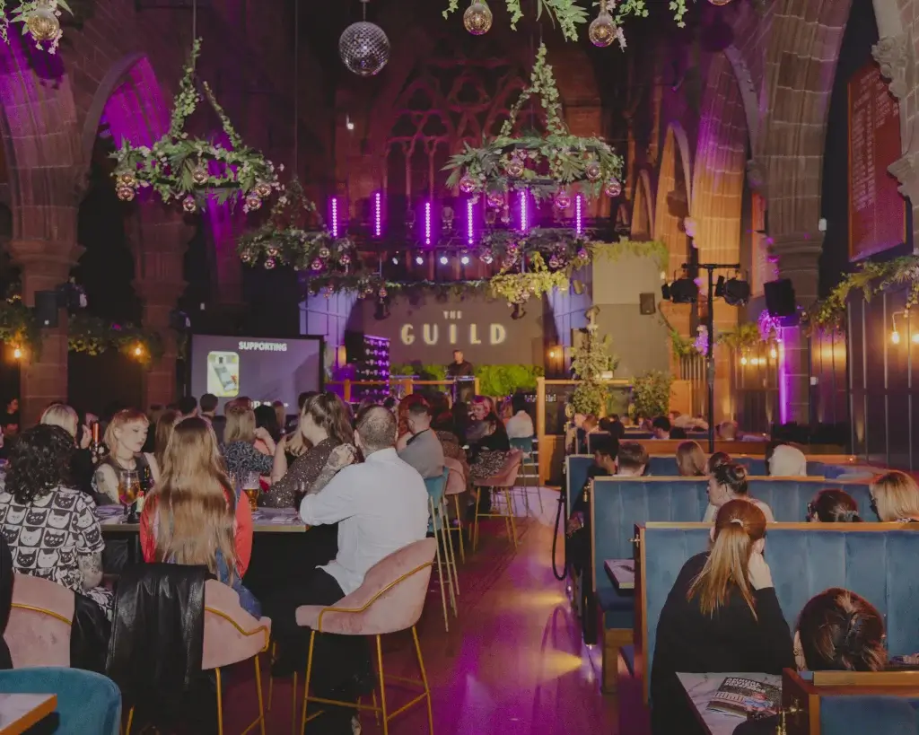 Crowded indoor event in a vaulted hall with hanging greenery and purple lights, diners seated facing a stage labelled 'GUILD'.