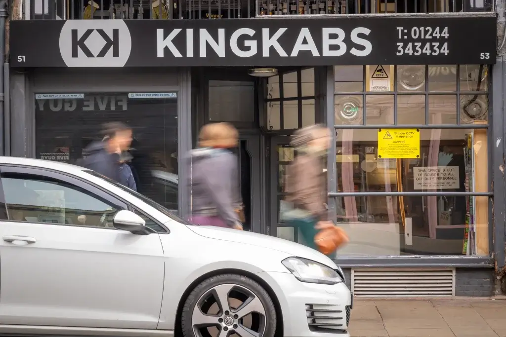 A white car parked in front of the KingKabs building, with blurred pedestrians walking past and a sign indicating CCTV in operation.
