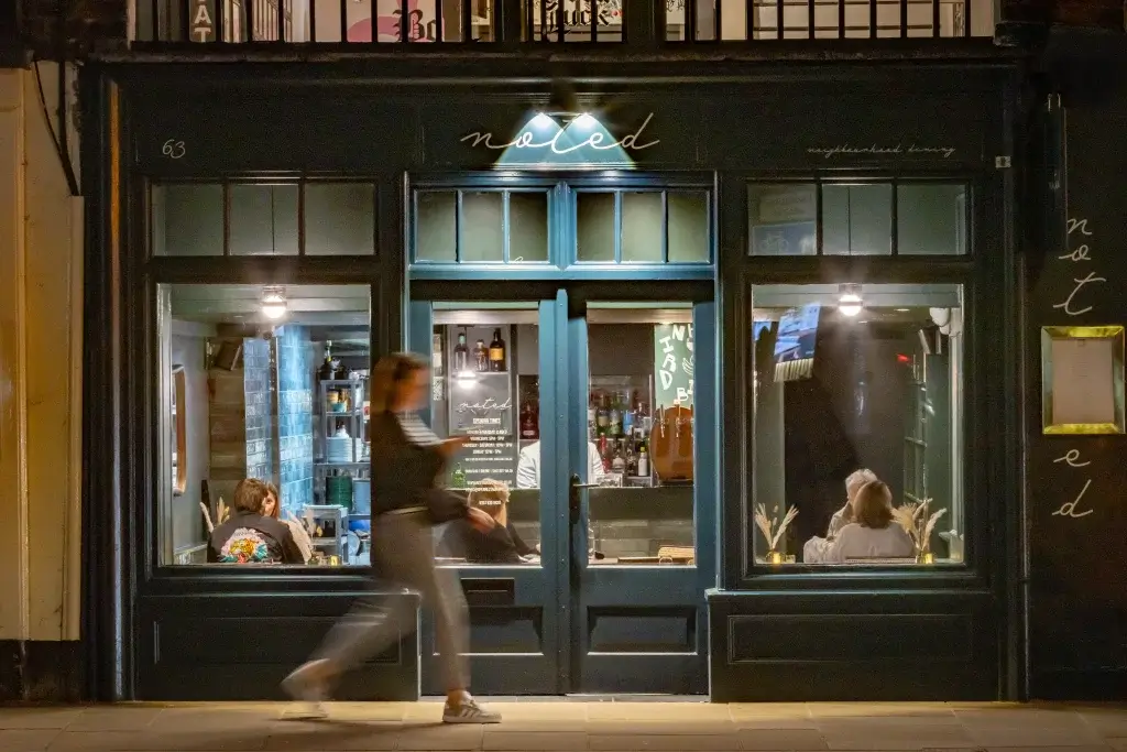 A dimly lit bar named "noted," with shelves stacked with bottles, showcasing a cozy atmosphere and patrons enjoying their evening.