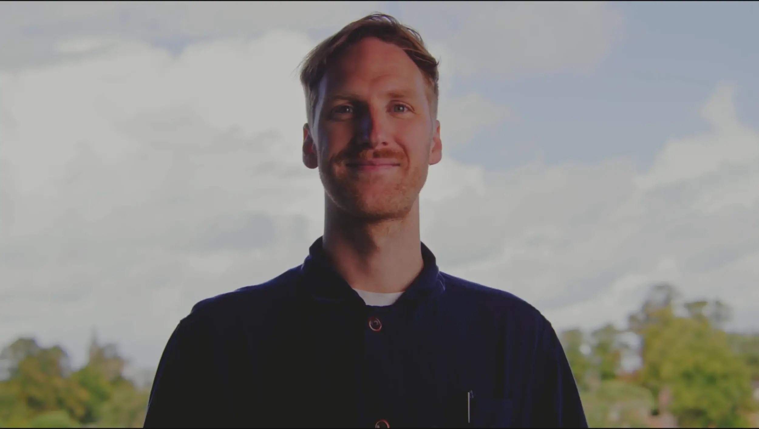 A man in a navy shirt stands against a backdrop of a bright sky with soft clouds and greenery in the distance.