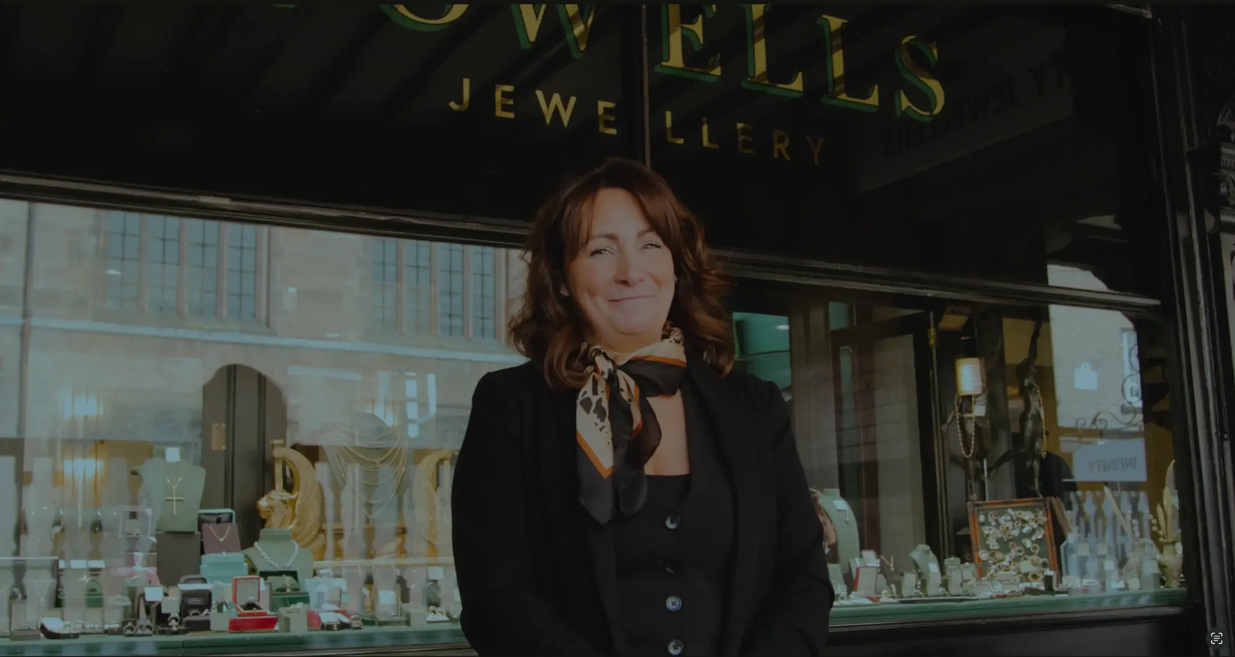 A woman stands in front of a jewellery store, showcasing an array of elegant jewellery displayed in the window behind her.