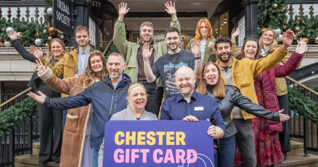 A group of diverse individuals stands on a staircase, celebrating with arms raised, holding a "Chester Gift Card" sign in front.